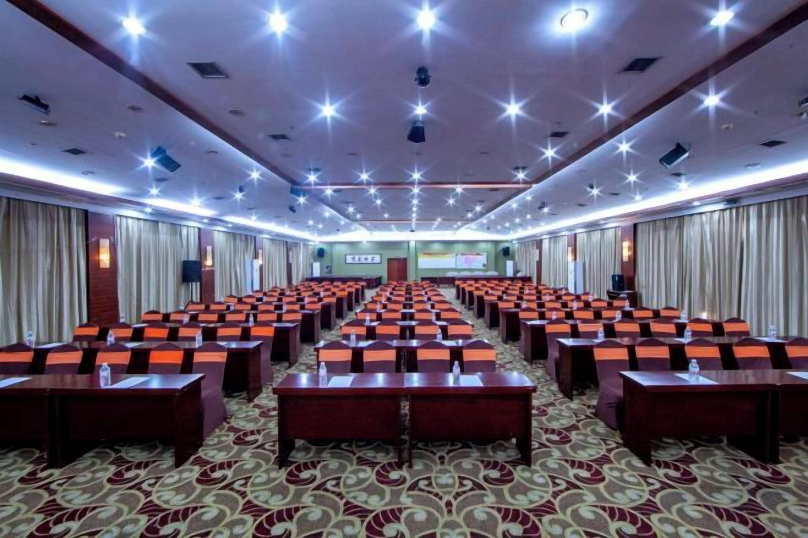 A conference room with rows of tables and chairs, a stage with a screen, and a carpeted floor.