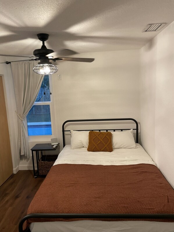 Un dormitorio con cama, ventilador de techo, ventana con cortinas y una mesita de noche con lámpara.