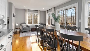 Een moderne keuken met houten vloeren, een eethoek met een lange tafel en stoelen, en grote ramen voorzien van gordijnen.