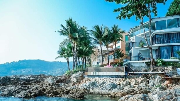 Una propiedad costera con costa rocosa, palmeras y un edificio moderno con balcones con vistas al mar.