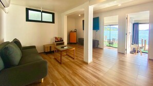Una sala moderna con sofá, mesa de centro y televisor. Cuenta con una zona de cocina con refrigerador y mesa de comedor. El piso es de madera y hay ventanas grandes que ofrecen vistas al mar.