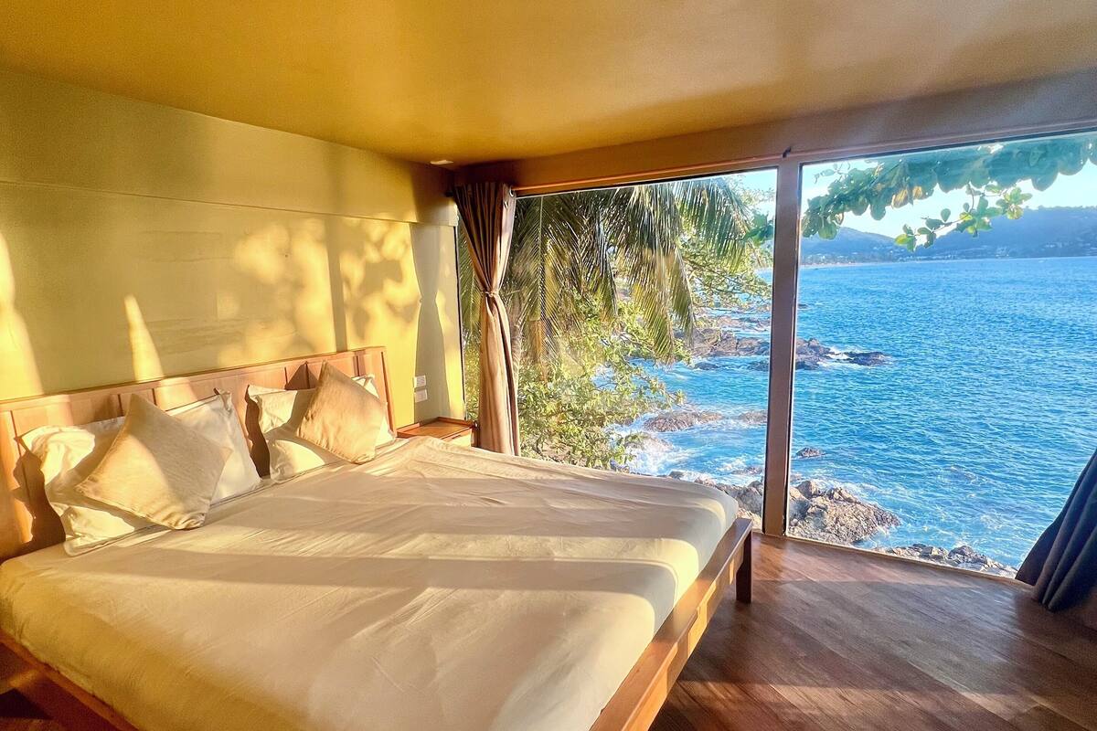 A bedroom with a large bed, wooden flooring, and a view of the sea through a large window.