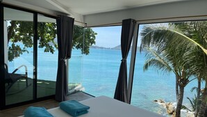 Un dormitorio con una cama grande, dos toallas azules, una silla y vista al mar a través de amplias ventanas.