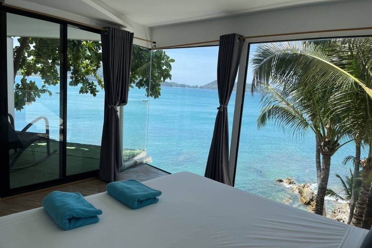 A bedroom with a large bed, two blue towels, a chair, and a view of the sea through large windows.
