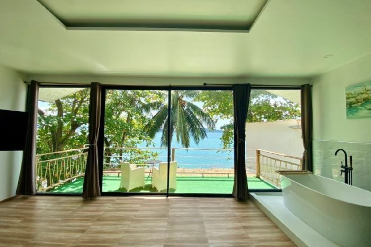 Quarto com uma grande porta de vidro deslizante que dá para uma varanda com vista para o mar e palmeiras.