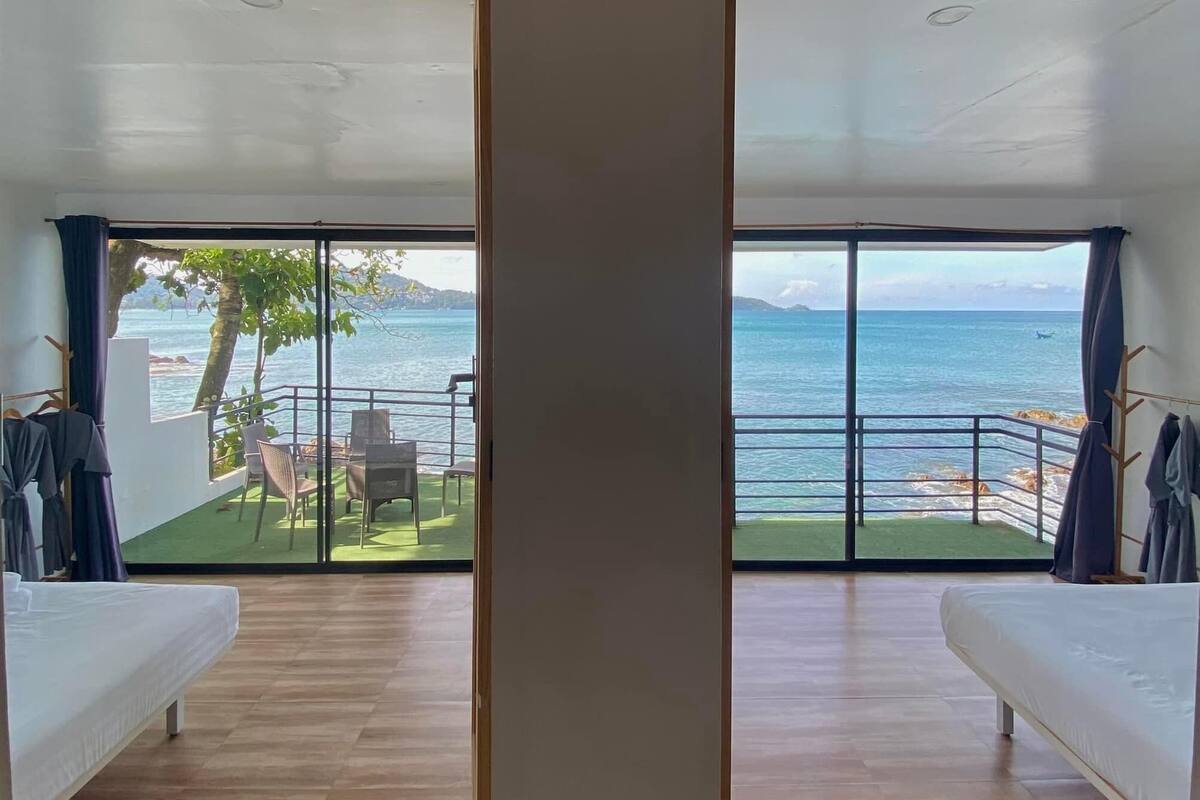 A room with two beds, large sliding glass doors leading to a balcony with a view of the sea and a grassy area with chairs.