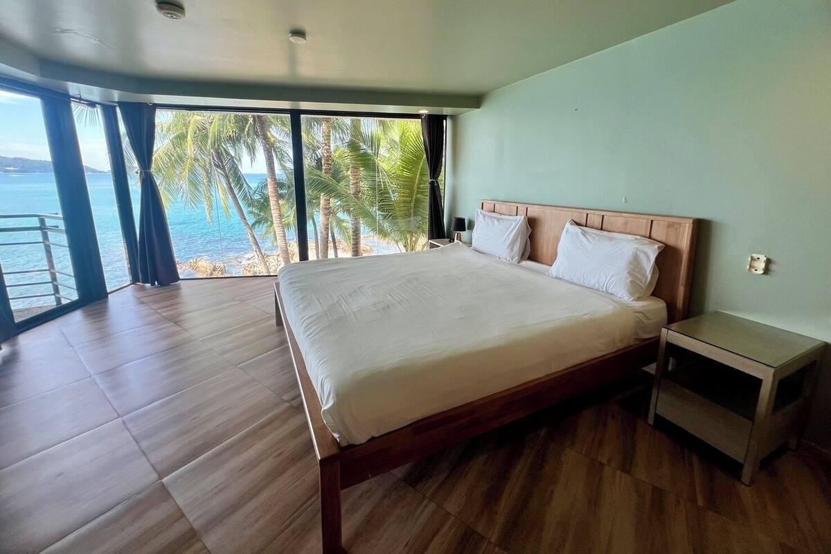 A bedroom with a large bed, a nightstand, and a view of the ocean through large windows.