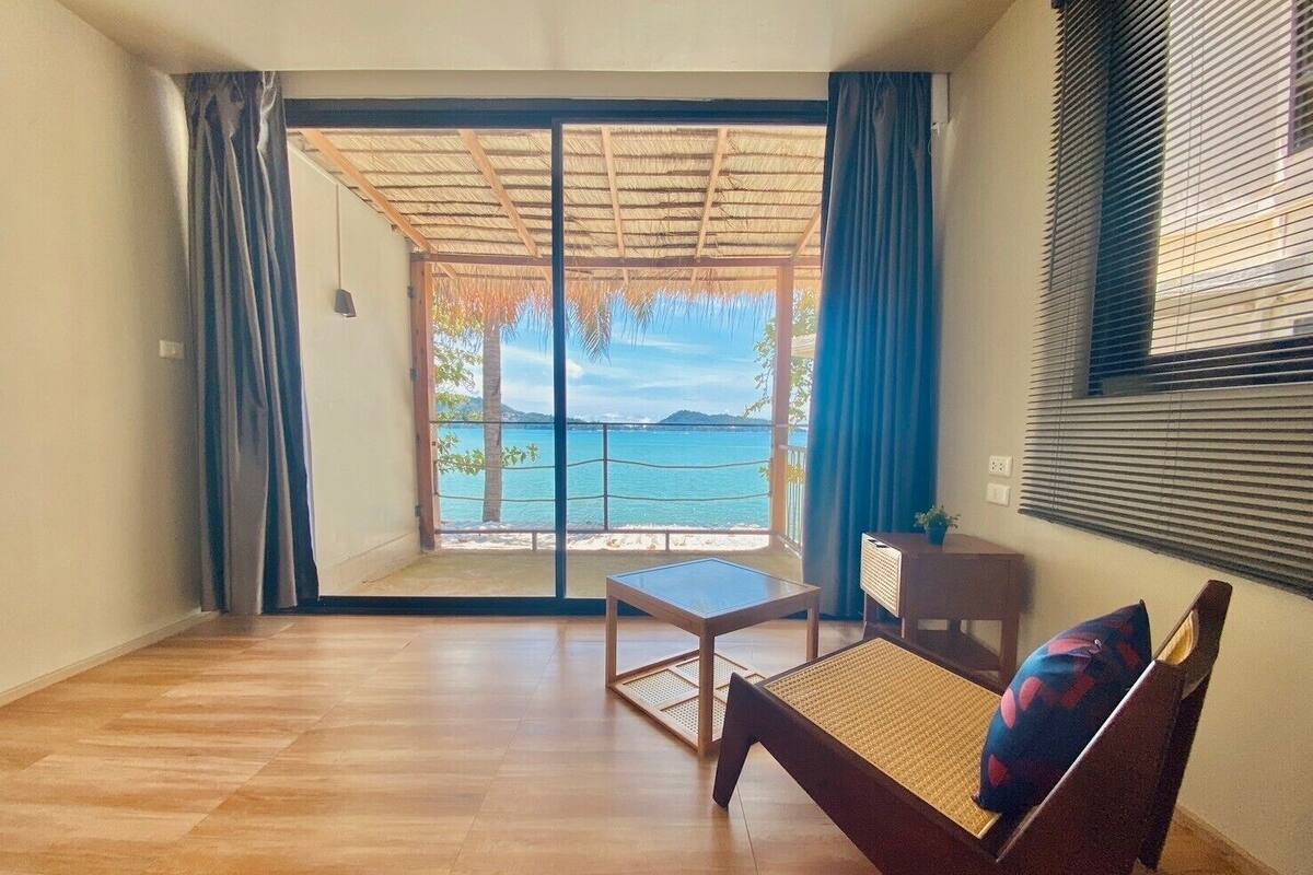 A room with a wooden floor, a sofa with a cushion, a small table, and a view of the sea through a glass door.