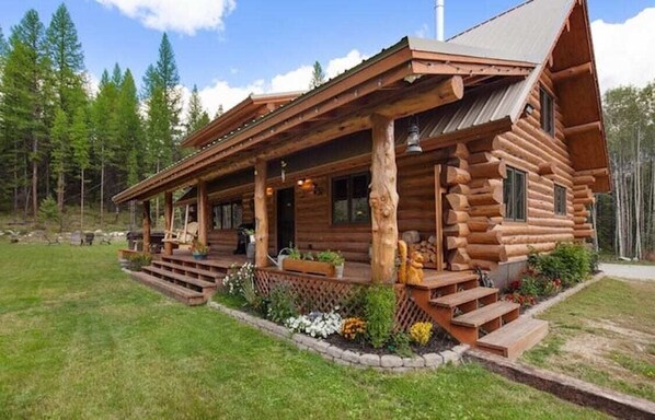 A log cabin with a porch, steps leading to the entrance, and a garden area with flowers and plants.