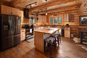 A log cabin kitchen with wooden cabinets, a stainless steel refrigerator, a central island with bar stools, and a large window.