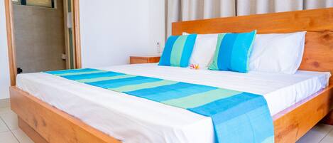 Una cama de madera con ropa de cama blanca y azul, una mesita de noche de madera y una ventana con cortinas.
