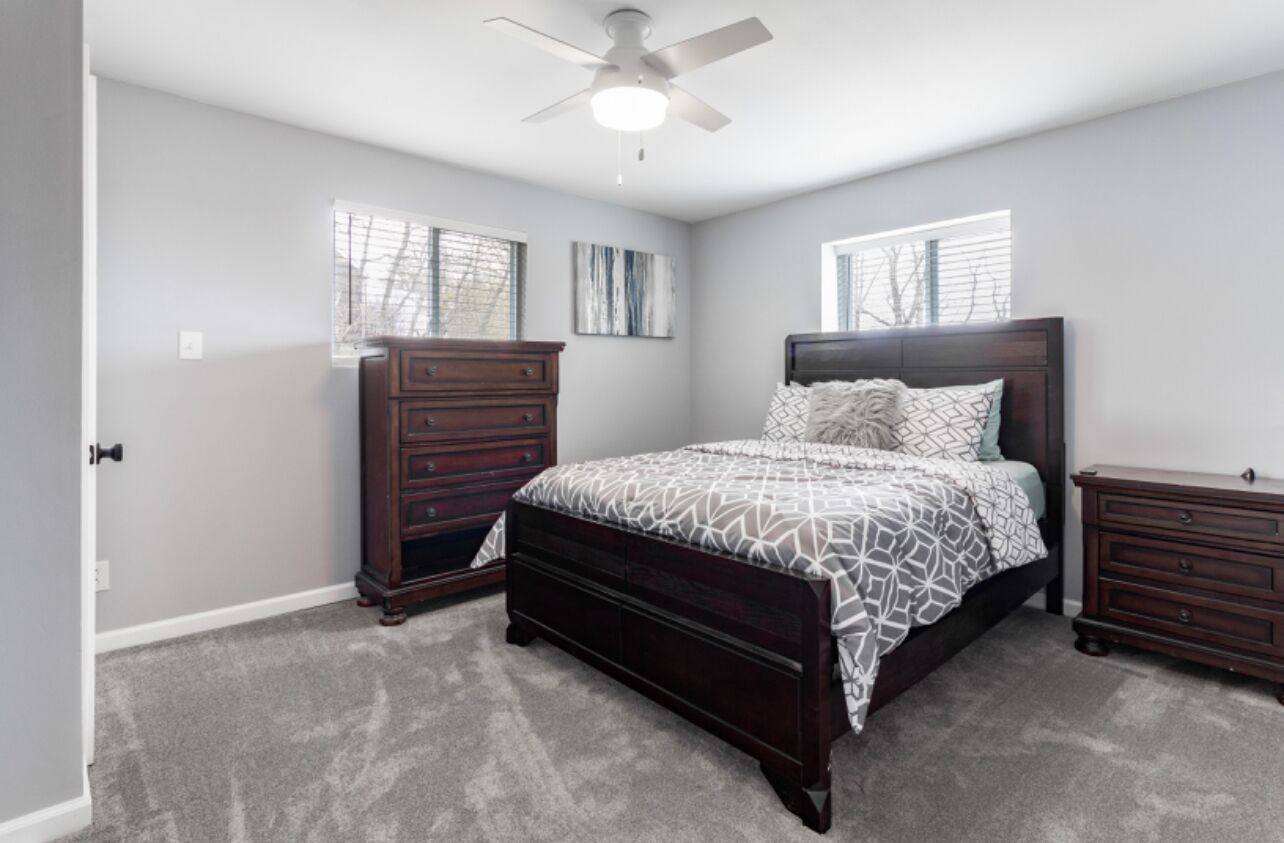 A bedroom with a bed, two nightstands, a dresser, a ceiling fan, and two windows with blinds.