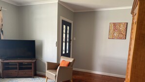 Una sala de estar con televisor de pantalla plana sobre un mueble de madera, un sillón color beige con un cojín naranja, una alfombra estampada y un aparador de madera.