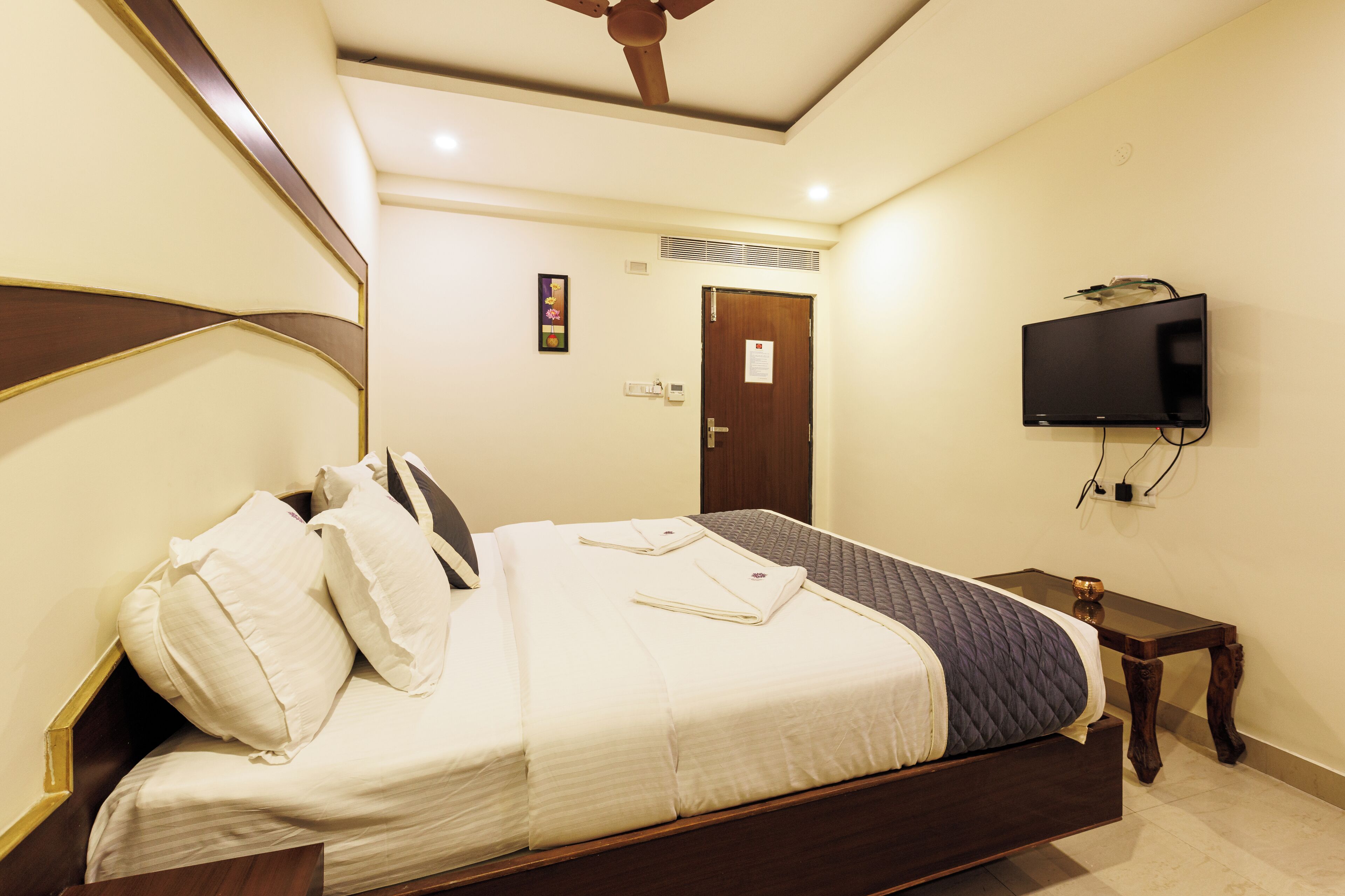 A neatly made bed with pillows, a bedside table, a television mounted on the wall, and a door in the background.