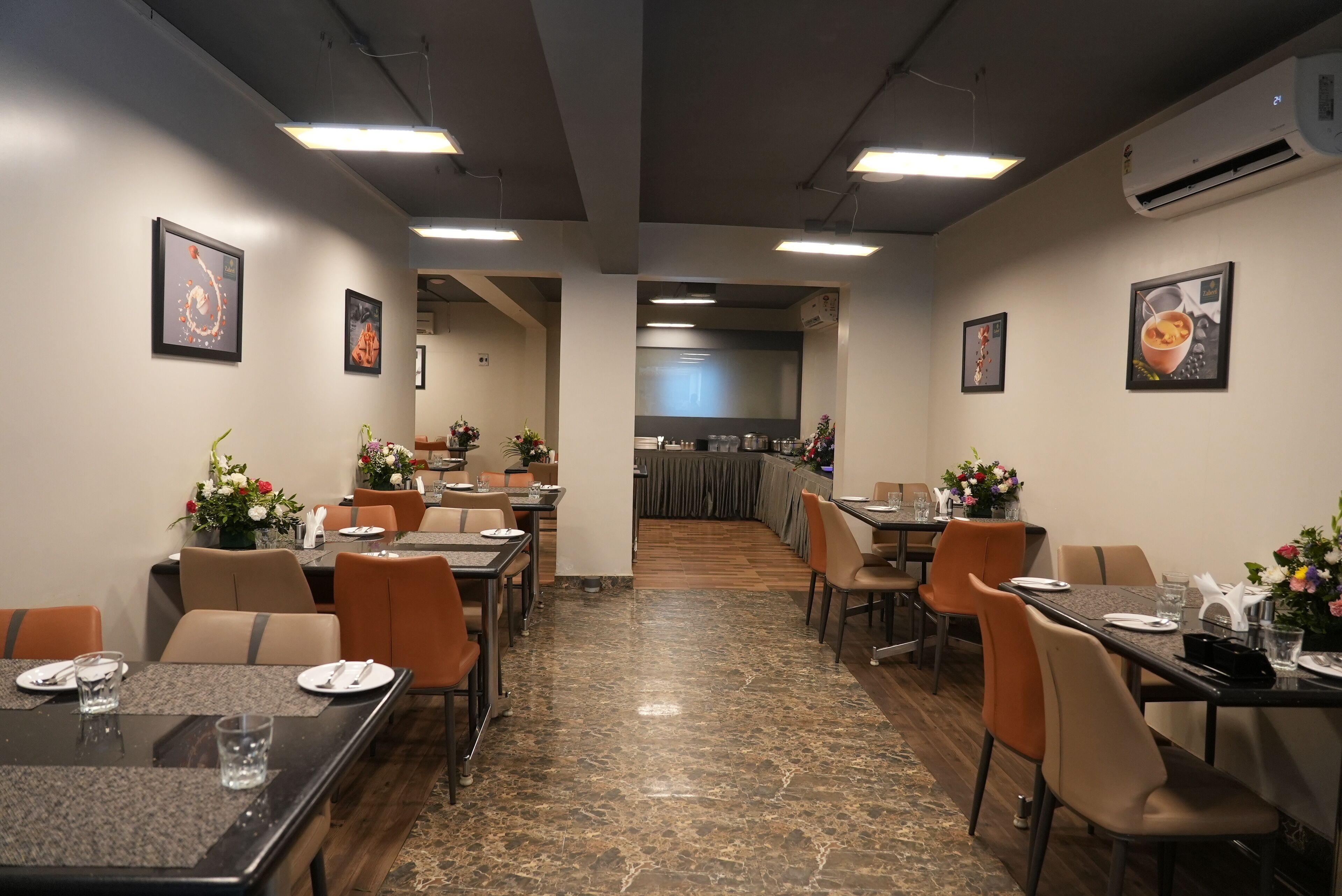 A restaurant with tables and chairs, a kitchen in the background, and framed pictures on the walls.