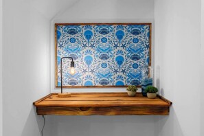 A wooden shelf with a blue patterned wall behind it, a floor lamp, and small potted plants.