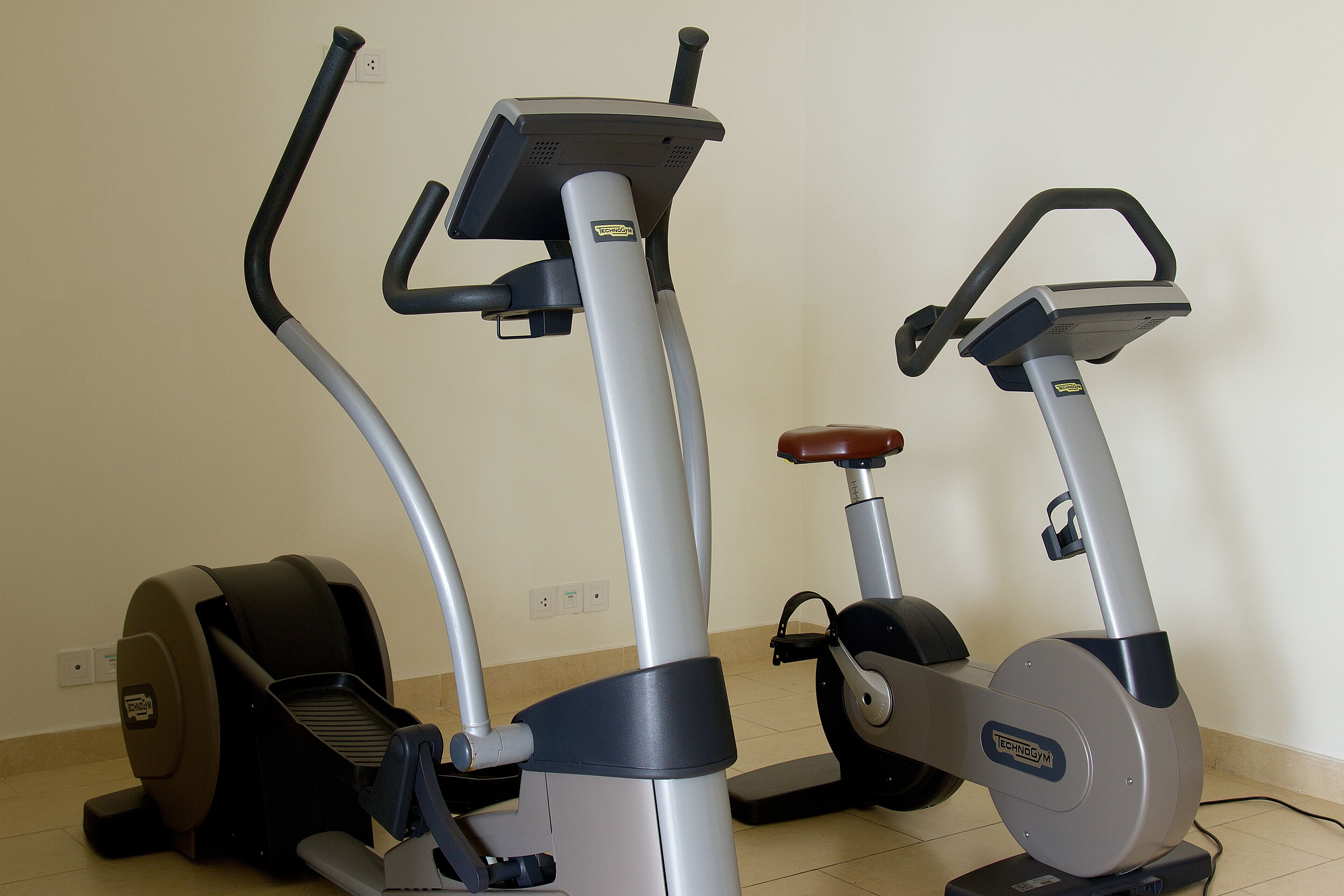 Two stationary exercise bikes in a room with a plain wall and a tiled floor.