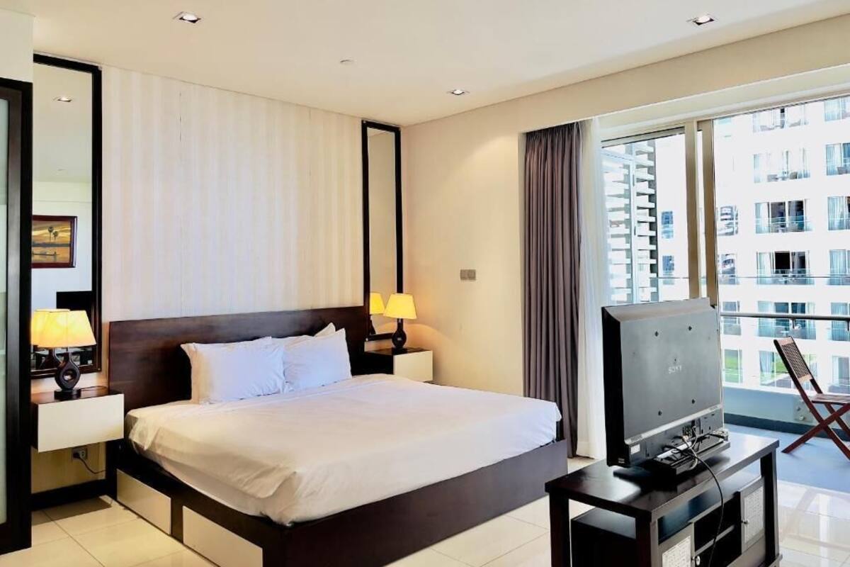 A hotel room with a large bed, a TV on a stand, a window with curtains, and a view of buildings outside.