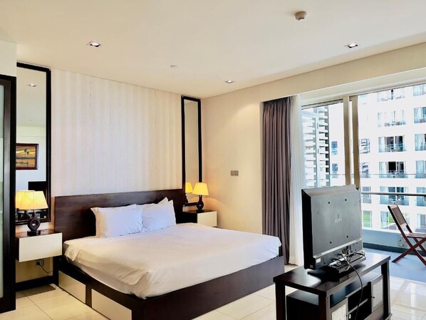 Habitación de hotel con una cama grande, un televisor sobre un mueble, una ventana con cortinas y vistas a edificios.
