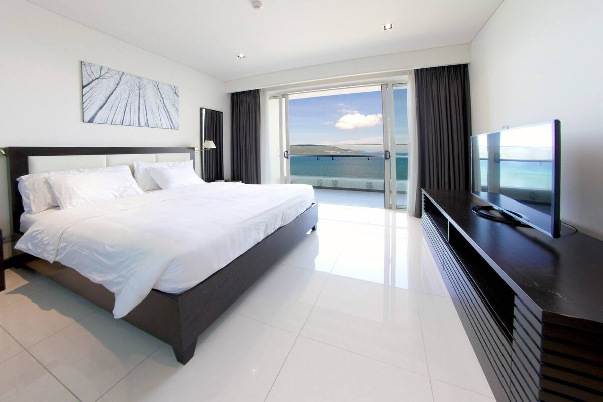 A modern hotel room with a large bed, a flat-screen TV, and a balcony with a view of the sea.