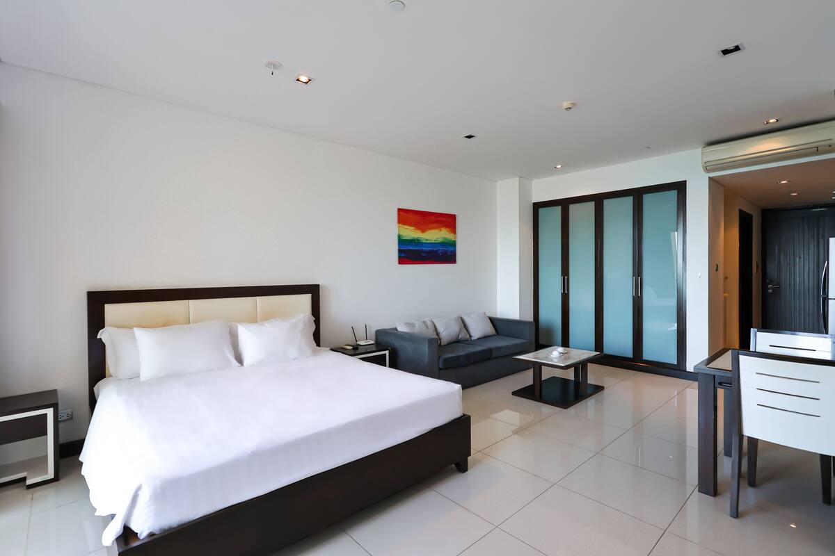 A modern hotel room with a large bed, a sofa, a desk, and a glass partition.