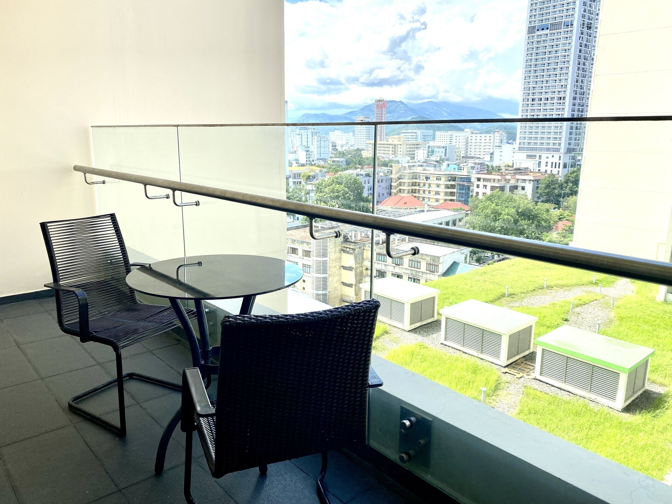 A balcony with a glass railing, two chairs, a small round table, and a view of a cityscape with buildings and greenery.