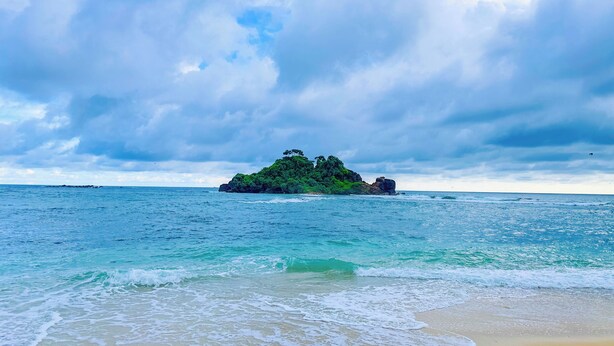 A beach with clear turquoise water, a sandy shore, and a small island with dense green vegetation in the distance. - The White House Villa (Ahangama)
