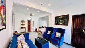 A spacious living area with a blue sofa set, a coffee table, and a dining table with chairs. There is a painting on the wall and a wooden cabinet with a clock. - The White House Villa (Ahangama)