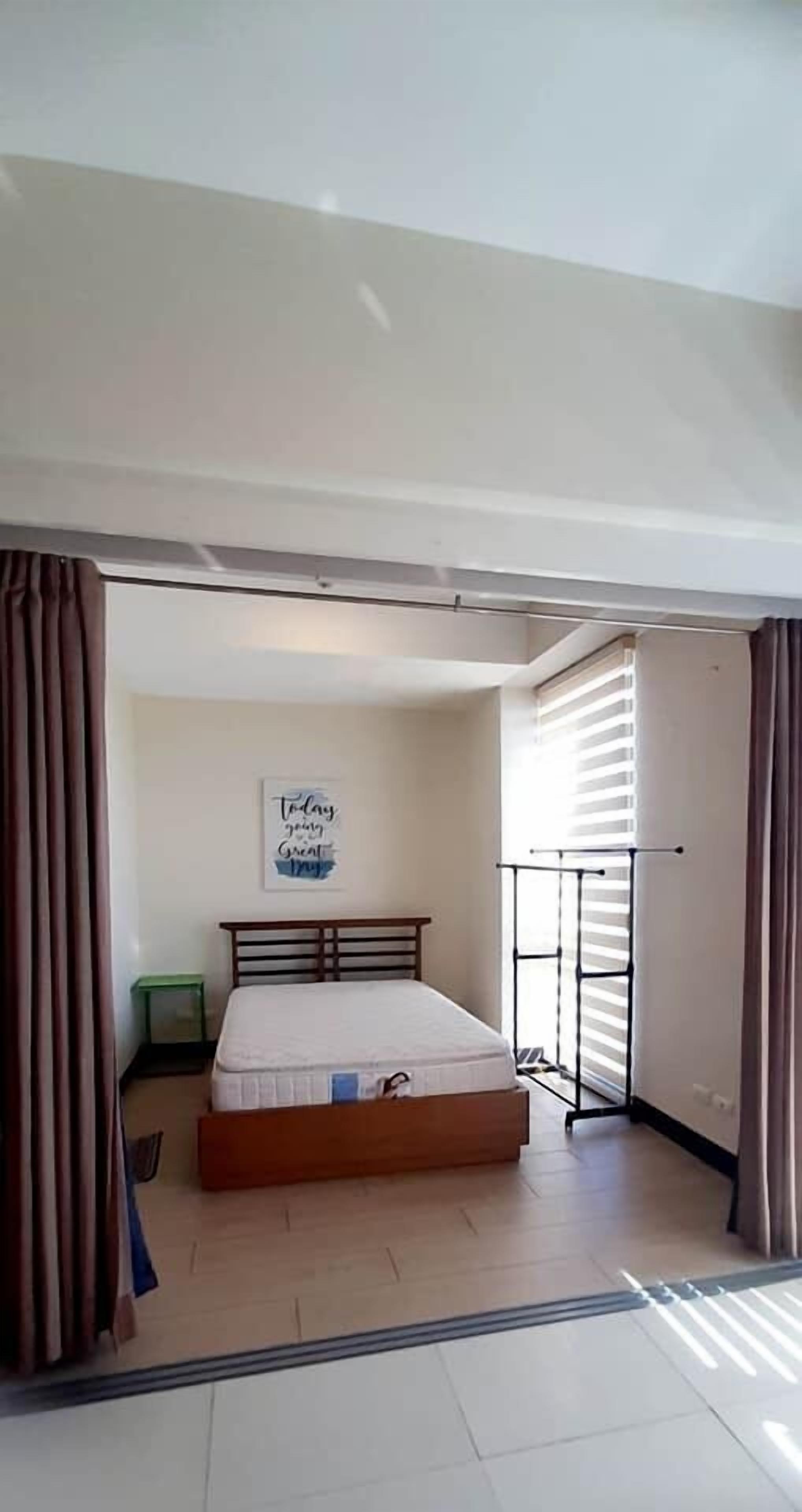 A bedroom with a bed, a small green stool, a wall with a motivational quote, and a window with blinds.