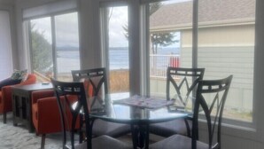 A dining area with a glass table surrounded by chairs, large windows offering a view of the outdoors, and a ceiling light fixture.