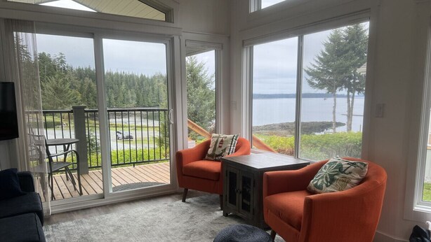 A cozy room with a large sliding glass door leading to a deck, a comfortable seating area with two armchairs and a small table, and a view of a lake and forest.