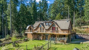 A large wooden house with multiple levels and a balcony, surrounded by a forested area.