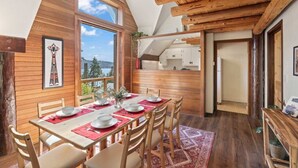 A dining area with a wooden table set for four, a view of the outdoors, and a kitchen area visible through an open door.