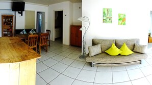 A living room with a wooden dining table, a sofa with yellow cushions, a floor lamp, and a TV mounted on the wall. - 80sqm two-room apartment - Wipfelder Montuerzimmer (Wipfeld)
