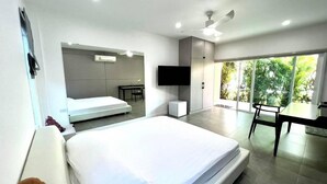 A modern hotel room with a large bed, a desk, a chair, a flat-screen TV, and a balcony with a view of greenery.