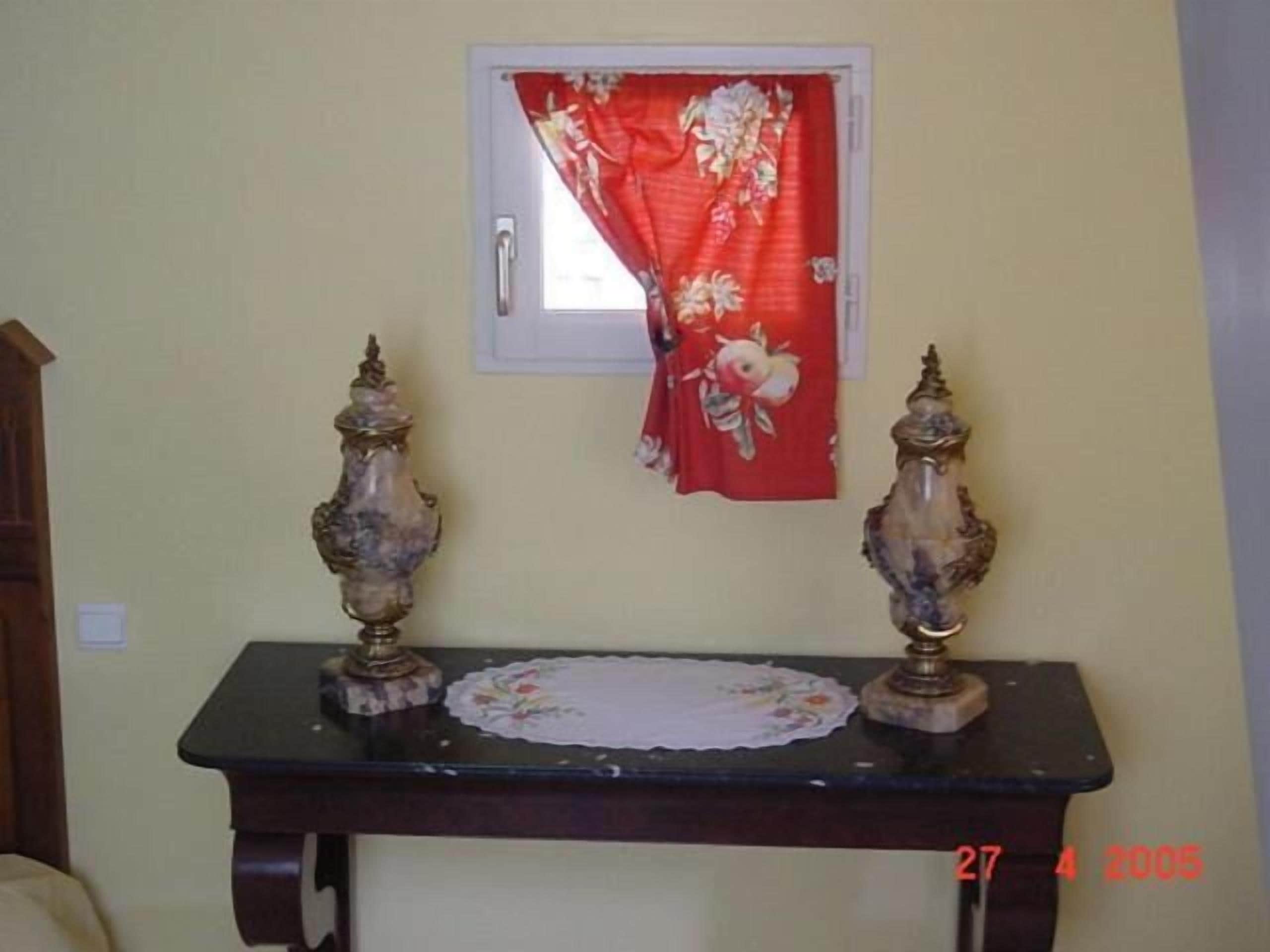 Una habitación con un ventanal pequeño cubierto por una cortina floral roja, una mesa de madera oscura con dos jarrones ornamentales y un mantel individual blanco sobre la mesa.
