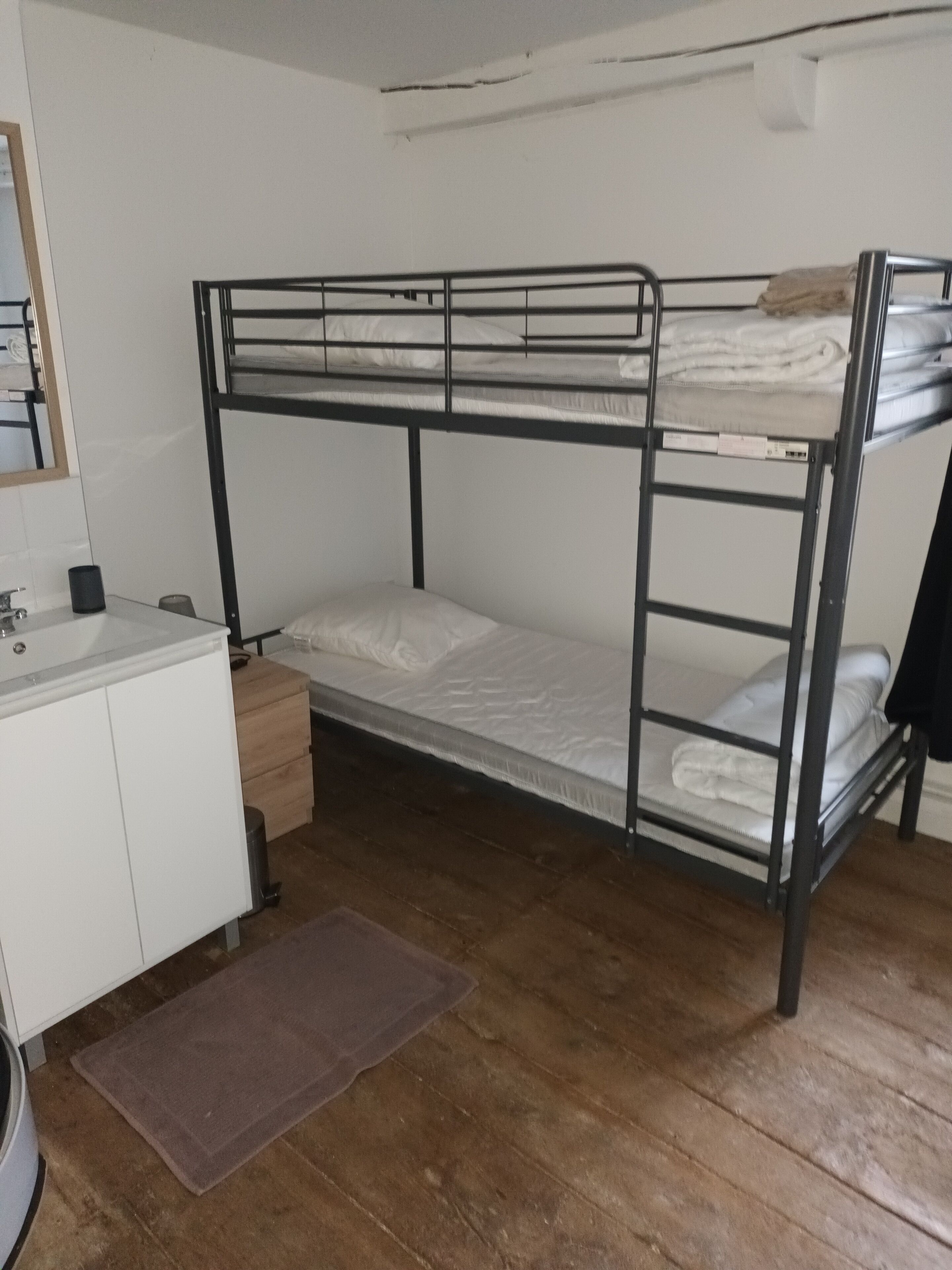 A bunk bed with a white mattress, a wooden floor, a small desk with a chair, and a mirror on the wall.