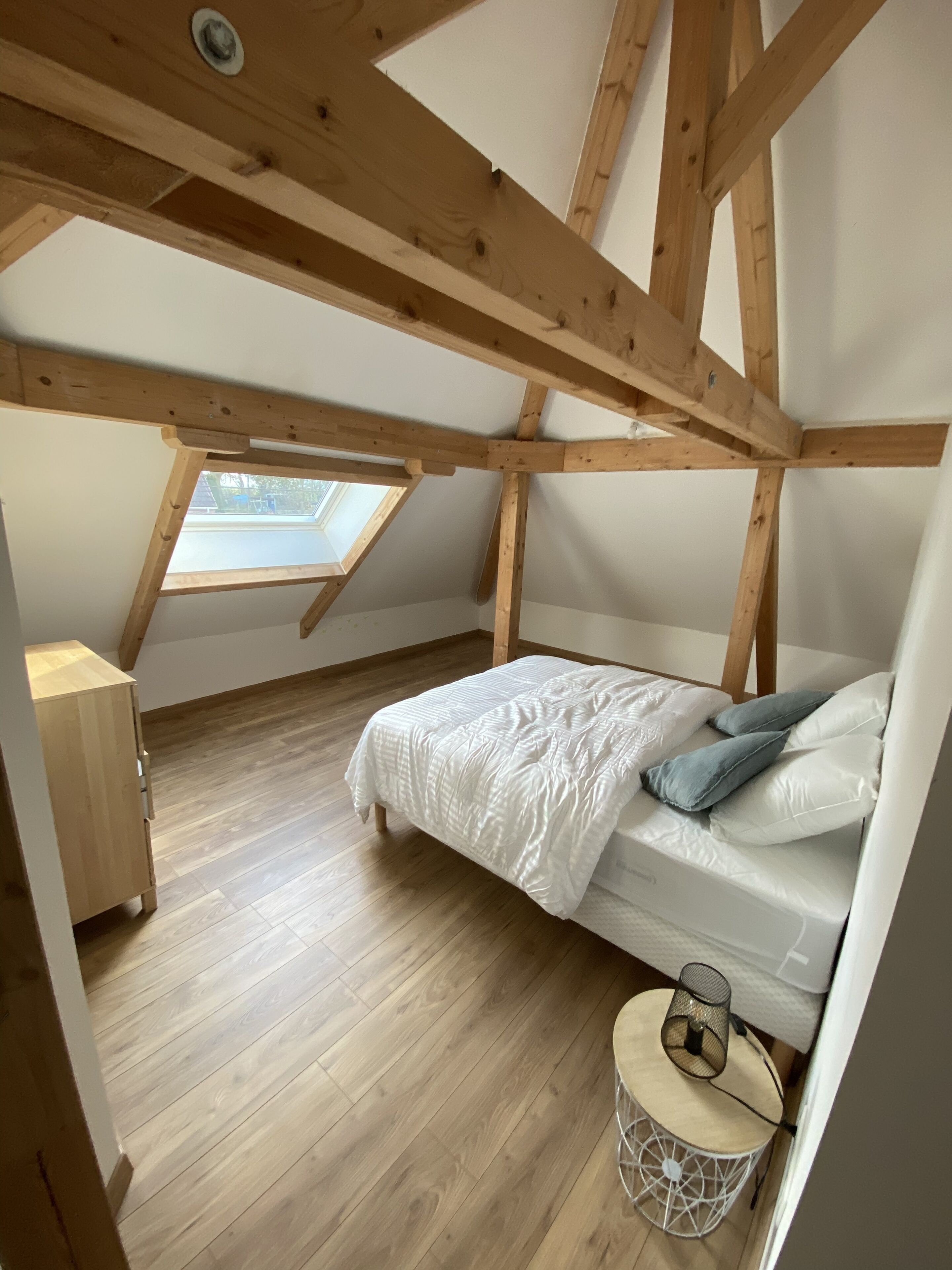 A bedroom with a sloped ceiling, wooden beams, a bed with white bedding, a small round table with a lamp, and a view through a skylight.