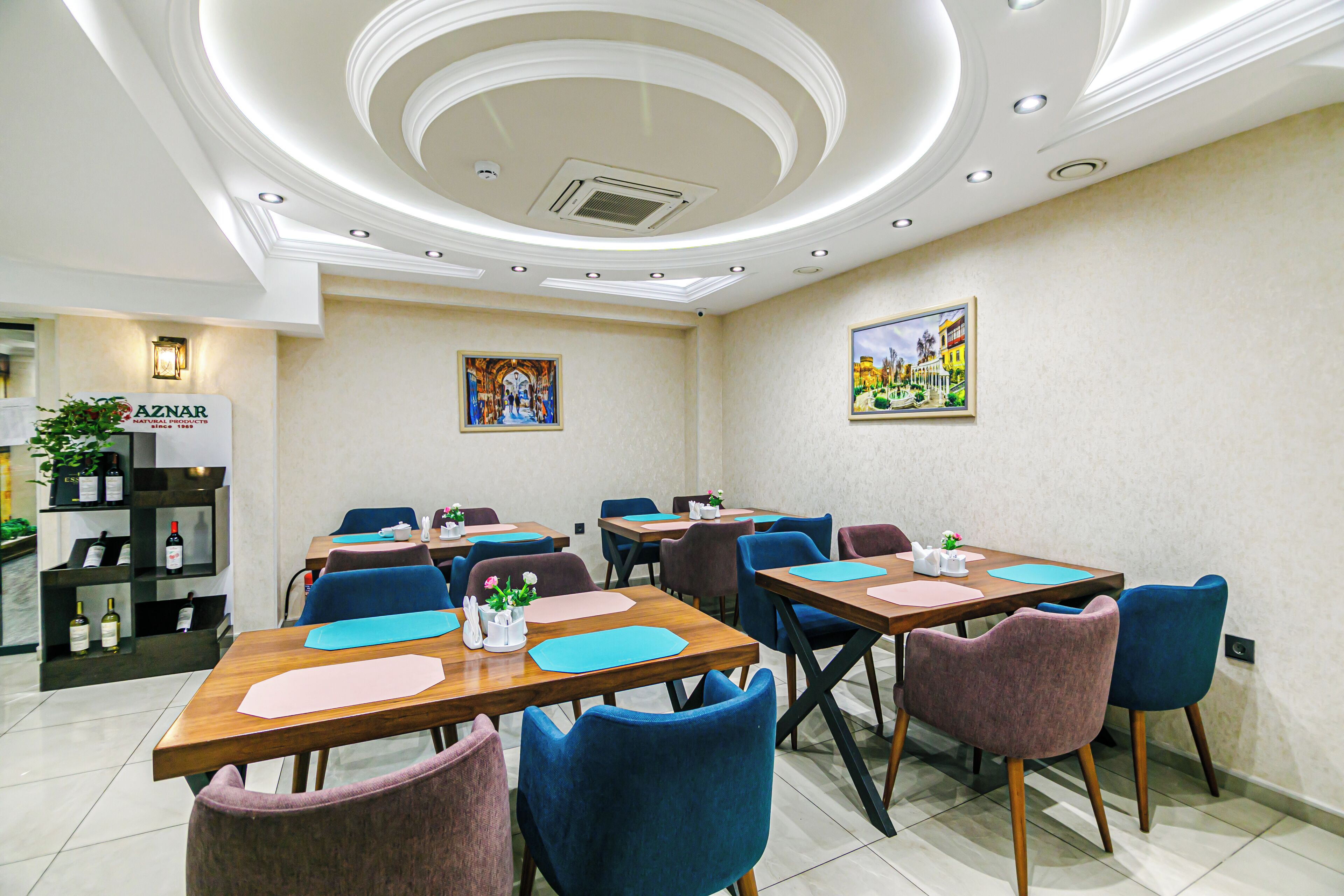 A hotel dining area with multiple tables and chairs, a ceiling with recessed lighting, and wall-mounted artwork.