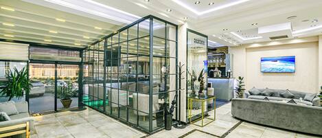 A modern hotel lobby with a glass partition, a reception desk, a sofa, and a large window overlooking the street.
