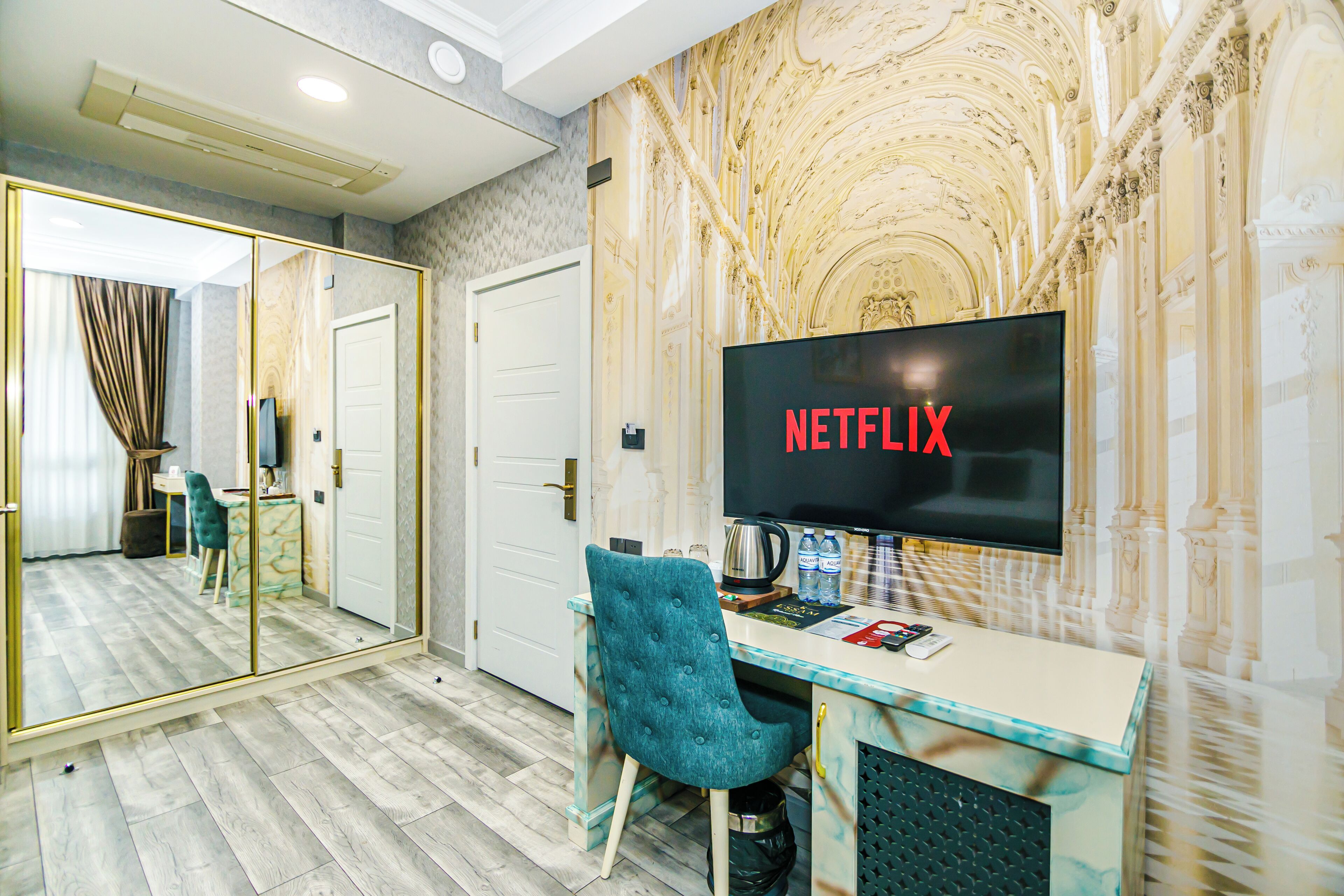 Ein modernes Hotelzimmer mit einem großen Spiegel, einem Schreibtisch mit Fernseher, der "NETFLIX" anzeigt, einem türkisfarbenen Sessel und einer Wand mit Marmoroptik.