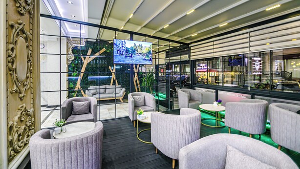 A modern lounge area with comfortable seating, a large TV screen, and a view of the cityscape through large windows. - Essam Deluxe Hotel (Baku)