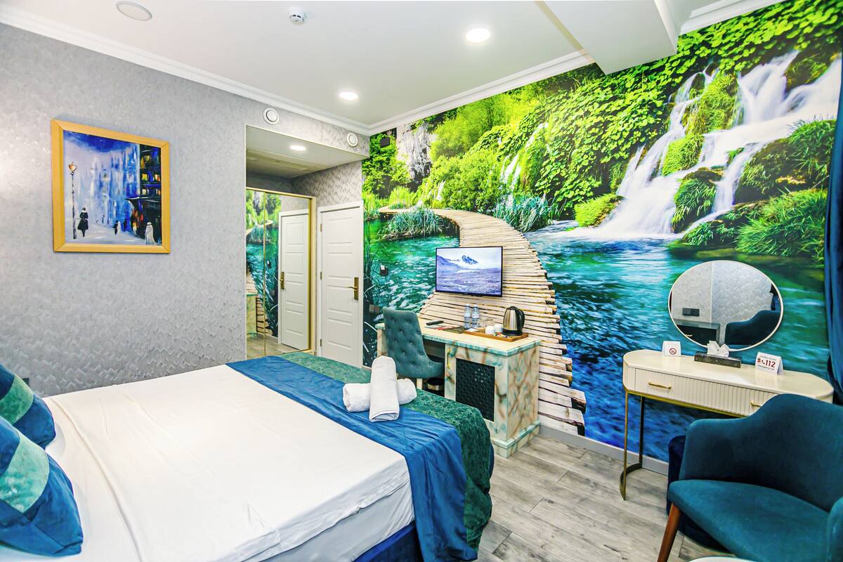 A hotel room with a bed, a desk with a computer, a chair, a vanity with a mirror, and a large mural of a waterfall.
