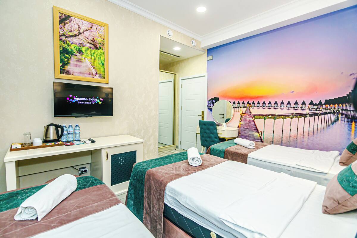A hotel room with two beds, a desk with a flat-screen TV, a painting of a sunset, and a view of a pier.