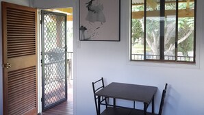 Um quarto com porta de madeira, uma mesa com cadeiras, uma obra de arte emoldurada e uma janela com vista para árvores.