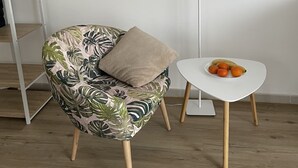 Huoneessa on kuviollinen nojatuoli, pieni pöytä, jossa on hedelmiä, lattiavalaisin ja seinää vasten nojaava tikapuu.