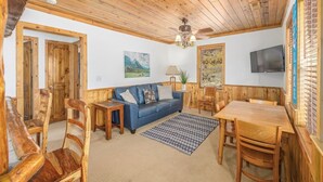 Una habitació acollidora amb sostre de fusta, un sofà blau, una taula de dinar de fusta i un televisor fixat a la paret.