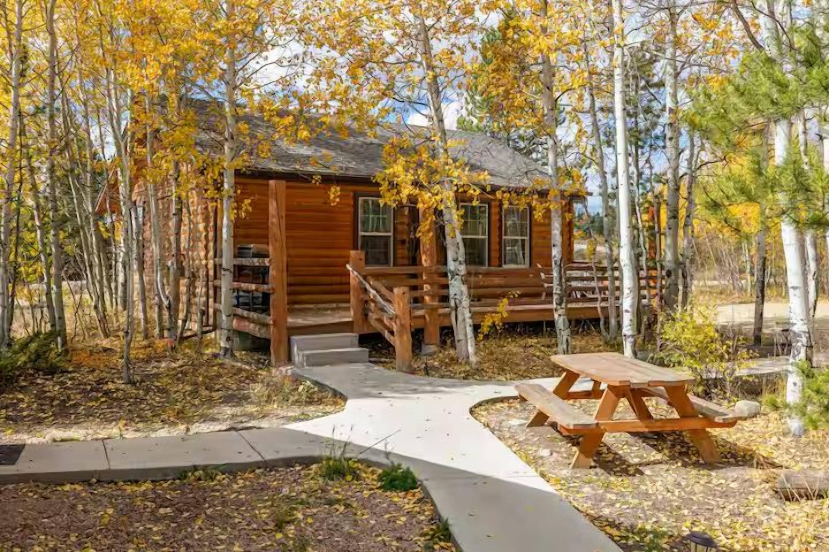 A wooden cabin with a porch, surrounded by trees with yellow leaves, and a picnic table in the foreground.