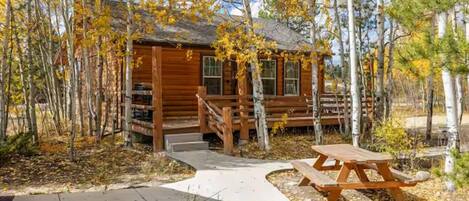A wooden cabin with a porch, surrounded by trees with yellow leaves, and a picnic table in the foreground.