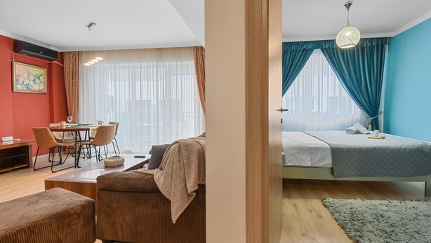 A modern hotel room with a dining area, a bed with a blue curtain, and a view of the city. - Sina Apartment (Voluntari)